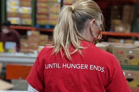 Food bank volunteer with pony tail and red shirt that says "Until Hunger Ends" on back